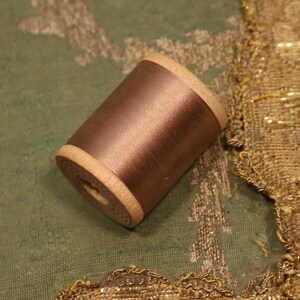May include: A wooden spool of metallic bronze thread with a smooth, satin-like finish. The spool features light-colored wooden ends with a circular design and a central hole, showcasing a cylindrical shape that holds the tightly wound bronze thread.