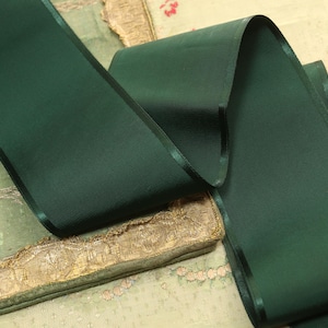 May include: A roll of dark green satin ribbon with a thin, darker green border. The ribbon is draped over a vintage fabric with gold trim and a faded floral pattern. The ribbon's texture is smooth and reflective.