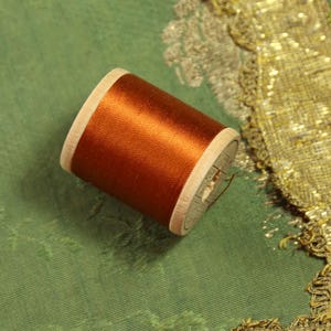 May include: A spool of metallic copper-colored thread wound around a light-colored wooden cylinder. The thread has a smooth, reflective sheen and is positioned at an angle, showing the circular ends of the wooden spool and the tightly wound fiber.
