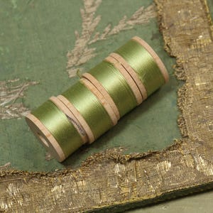 May include: A close-up of several spools of green thread. The thread is wound around wooden spools. The spools are arranged diagonally on a green fabric with gold trim. The fabric has a floral design.