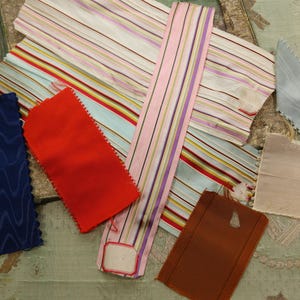 May include: Assortment of fabric scraps in various colors and patterns, including red, blue, pink, and brown. The fabrics are arranged on a light green surface.