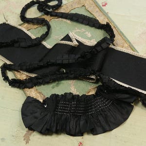 May include: Black fabric trim with white lace edging and a ruffled design. The trim is arranged on a vintage-style background with a gold-colored frame. The fabric has a satin-like sheen.