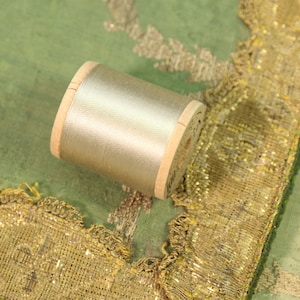 May include: A spool of silver thread rests on a green fabric with gold trim. The thread is wound around a wooden spool. The fabric has a gold embroidered border. The image showcases sewing and embroidery supplies.