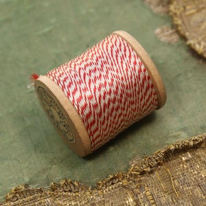 May include: A wooden spool of red and white twine. The twine is wrapped around the spool in a diagonal pattern. The spool is lying on a green surface with a gold and tan fabric in the background. The spool is a craft supply.