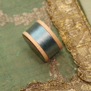 May include: A spool of teal blue thread wrapped in a wooden bobbin. The bobbin is lying on a green fabric with gold embroidery.