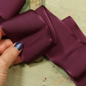 May include: Close-up of a deep plum-colored satin ribbon. The ribbon is folded and held by a hand with a dark blue nail polish. The edges of the ribbon are stitched. The ribbon is on a light green and gold surface.