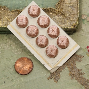 May include: A card of eight rose-colored, embroidered buttons with a cross-stitch design. A copper penny is next to the card for size comparison. The buttons are round and have a slightly raised center. The background is a light green fabric.
