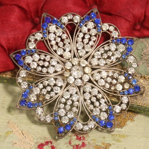 May include: A silver-toned brooch with a flower design. The flower is made of clear rhinestones and has blue rhinestones around the edges.