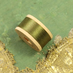 May include: A spool of olive green thread wound around a wooden bobbin. The bobbin has a white label with black text.