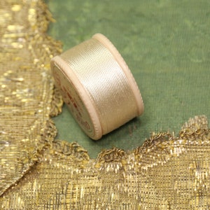 May include: A spool of pale gold thread rests on a green fabric, near a gold metallic trim. The thread is wound around a light-colored spool. The trim has a scalloped edge and a woven texture. The background is a muted green.