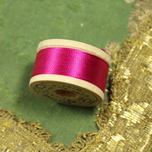 May include: A spool of bright pink thread wound on a wooden bobbin. The bobbin is labeled "100% Fine Silk Swiss Made" and "D.C.  Kreinik"