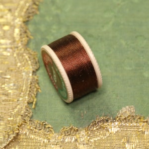 May include: A spool of brown thread with a white plastic bobbin. The thread is wound around the bobbin and is ready to be used for sewing or embroidery.