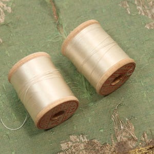 May include: Two spools of off-white thread sit on a green, textured surface. The spools have wooden sides and are filled with a fine, light-colored thread. One spool has a loose thread extending from it.