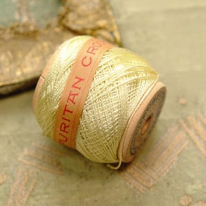 May include: A spool of pale yellow crochet thread wrapped around a wooden core. A tan paper label is wrapped around the center of the thread, featuring the red printed text PURITAN CROCHET. The spool has circular wooden ends with dark markings.