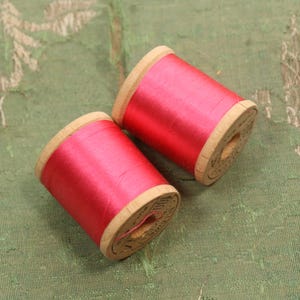 May include: Two spools of red thread with wooden sides sit on a green patterned surface. The thread is a vibrant red color, and the wooden spools have a natural wood grain. The spools are likely for sewing or crafting.