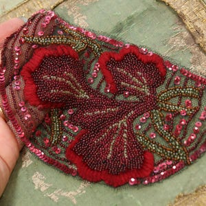 May include: A decorative applique featuring a floral design. The applique is primarily burgundy with red embroidered flowers and green stems. It is embellished with sequins and beads, and is set against a sheer, dark mesh.