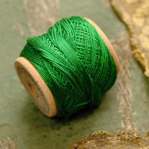 May include: A wooden spool wrapped with vibrant emerald green embroidery thread. The spool features a light tan wood grain finish on the circular ends, with a visible central hole and dark printed text along the edge of the wooden spool face.