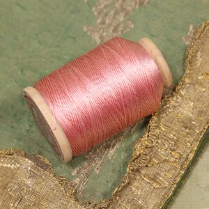 May include: A spool of pink thread wound around a wooden bobbin. The thread is smooth and shiny, and the bobbin is a light brown color.