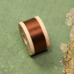 May include: A spool of brown thread wound around a wooden bobbin. The bobbin is labeled with text.