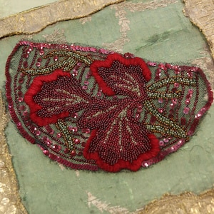 May include: A close-up of a decorative textile piece featuring a floral design. The piece is adorned with red beads, sequins, and embroidery on a green mesh background. The edges are trimmed with gold and a metallic border.