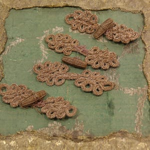 May include: Close-up of five beige frog closures, each with a looped design and a cylindrical toggle. They are arranged on a textured, light green fabric with a gold border. These closures are likely for clothing or decorative purposes.