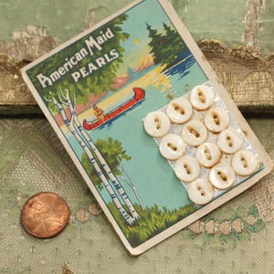 May include: Vintage card of "American Maid Pearls" buttons. The card features a scenic illustration of a canoe on a lake. Twelve cream-colored, round buttons are attached. A penny is in the lower left corner.
