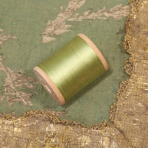 May include: A spool of green thread wound around a wooden bobbin. The bobbin is sitting on a green fabric with gold trim.