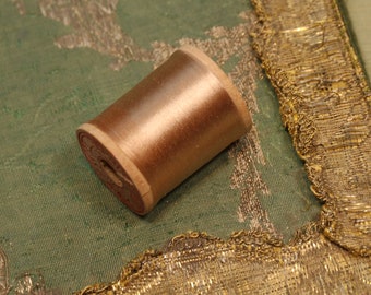 May include: A spool of brown thread wound around a wooden bobbin. The bobbin has a red label with white text. The spool is sitting on a green fabric with a gold trim.