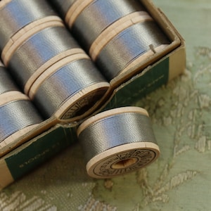 May include: A box of silver-gray thread spools. The spools are made of wood and are filled with a metallic-looking thread. The box is made of cardboard and has a green label. The spools are arranged neatly inside the box.