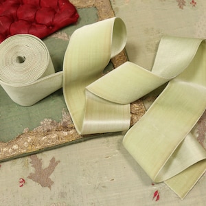 May include: A roll of pale green ribbon with a velvety texture, unrolled to reveal its satin sheen. The ribbon is displayed on a distressed green surface with gold trim and a red, tufted cushion in the background. Decorative craft supply.