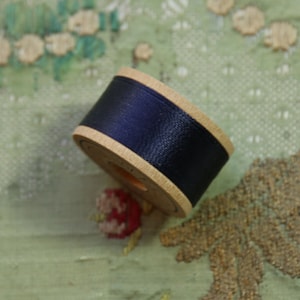 May include: A wooden spool wrapped with dark navy blue ribbon. The spool has a light tan natural wood finish with visible grain. The ribbon is smooth, opaque, and tightly wound around the center of the spool, creating a cylindrical shape.