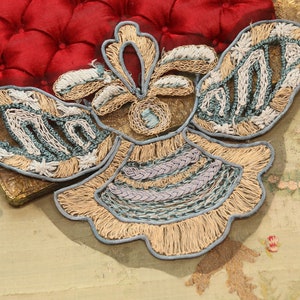 May include: A beige and blue embroidered applique in the shape of a butterfly. The applique has a detailed design with intricate stitching and a fringed edge.