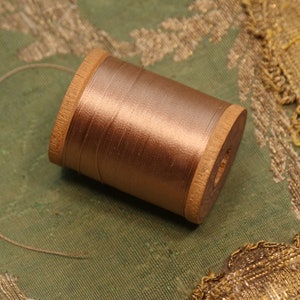 May include: A spool of copper-colored thread sits on a green fabric with gold embroidery. The thread is wound around a wooden spool, with a strand extending outwards. The background features detailed gold embellishments, suggesting a craft or textile project.