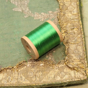 May include: A spool of bright green thread wound around a wooden bobbin. The spool is sitting on a green fabric with gold trim.