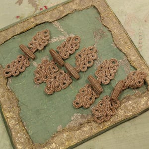 May include: Close-up of a collection of antique tan-colored textile closures, each with a looped end and a cylindrical toggle. They are arranged on a square, light green fabric with a gold border.