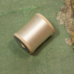 May include: A spool of white thread wound around a wooden bobbin. The bobbin has a blue and white label.