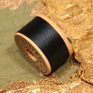 May include: A spool of black thread wound around a wooden bobbin with a label that reads "SERA DE LIN"
