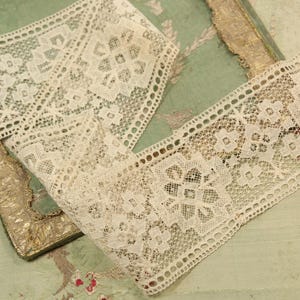 May include: Close-up of cream-colored lace ribbon with a floral and geometric pattern. The lace is laid out on a vintage-style background with a gold-trimmed edge. The ribbon has a delicate, open weave.