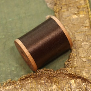 May include: A spool of dark brown thread with a wooden bobbin. The thread is wound tightly around the bobbin and has a smooth, shiny finish.