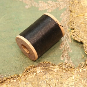 May include: A wooden spool of black sewing thread with a cylindrical shape. The spool features light-colored wood ends, with one end showing a circular paper label with printed text. The thread is wound tightly around the central wooden core.