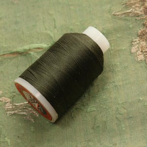 May include: A spool of dark green thread with a white base and a red and white label. The thread is tightly wound around the spool. The background is a textured, faded green fabric.