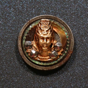 May include: A circular, gold-toned button with a raised image of a woman's face wearing a crown. The button is surrounded by a green and silver border and has small, silver-toned studs around the edge.