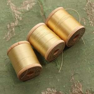 May include: Three spools of golden yellow silk thread on a green patterned fabric. The wooden spools have a light brown color and are labeled with text. The silk thread is tightly wound around the spools, creating a smooth, shiny surface.