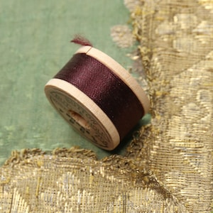 May include: A wooden spool of dark burgundy thread, with a small tuft of thread extending from the top. The spool is set against a backdrop of green fabric and gold trim, suggesting a sewing or craft theme.