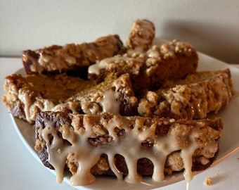 May include: A plate of sliced pumpkin bread with a crumb topping and a drizzle of light-colored glaze. The bread is a warm brown color, and the glaze is dripping down the sides. The plate is white, and the background is a neutral color.