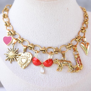 May include: Gold-coloured charm necklace featuring a variety of pendants, such as a pink heart, sun, heart-shaped locket, red bra, leopard, and cowboy boot. The necklace is displayed on a white mannequin.
