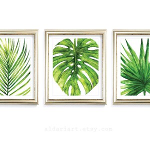 Puede incluir: Tres impresiones enmarcadas en acuarela con vibrantes hojas tropicales verdes. La obra de arte incluye una hoja de palma, una hoja de monstera y una hoja similar a un helecho, todo sobre un fondo blanco. Los marcos tienen un acabado dorado. El nombre de la tienda Etsy es visible.