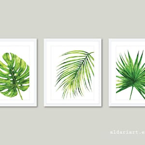 Puede incluir: Tres impresiones enmarcadas en acuarela con hojas tropicales verdes. Las impresiones incluyen una hoja de monstera, una fronda de palmera y una hoja de palma abanico, sobre un fondo blanco. Los marcos son blancos. El nombre de la tienda Etsy es visible en la parte inferior.