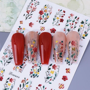 May include: A sheet of nail art stickers featuring floral designs and the word "Forever". The stickers include red, yellow, and green flowers and leaves. Five nails are shown with red and floral designs.