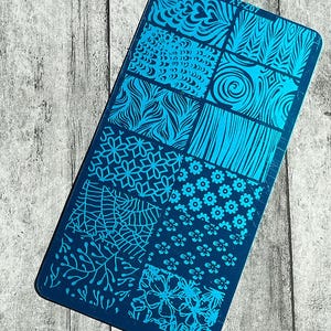 May include: A rectangular, navy blue nail art stamping plate with various patterns in shades of blue. Designs include floral, geometric, and abstract shapes. The plate is set against a weathered, gray wooden surface.